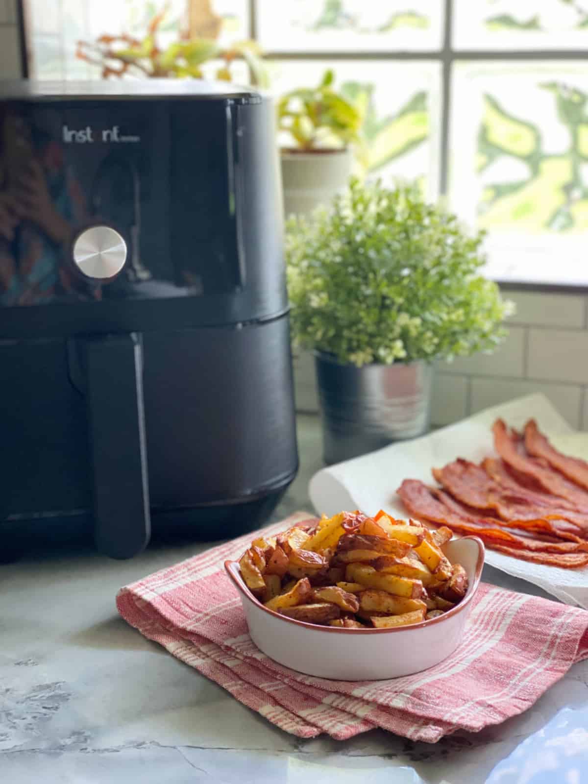 Fried potatoes in white dish with air fryer and cooked bacon in the background