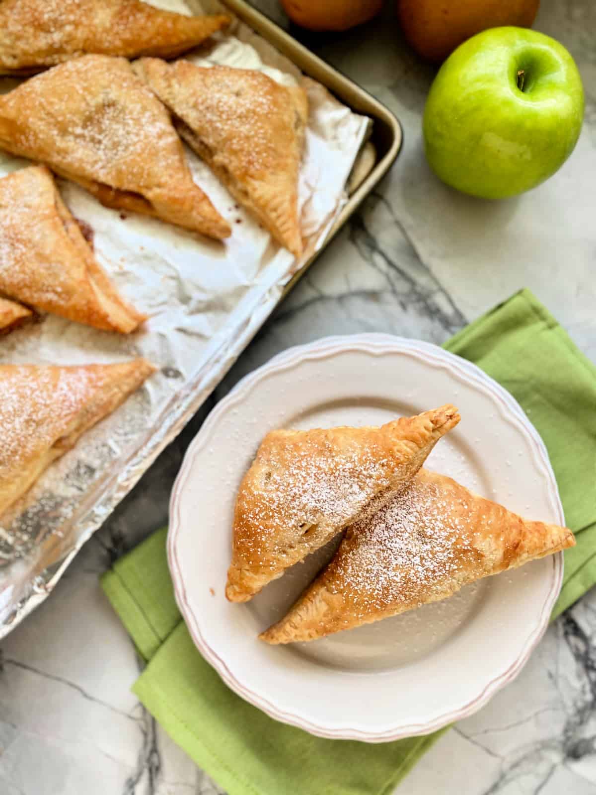 Top view of two turnovers on a white plate with a baking tray filled with other turnovers next to it.