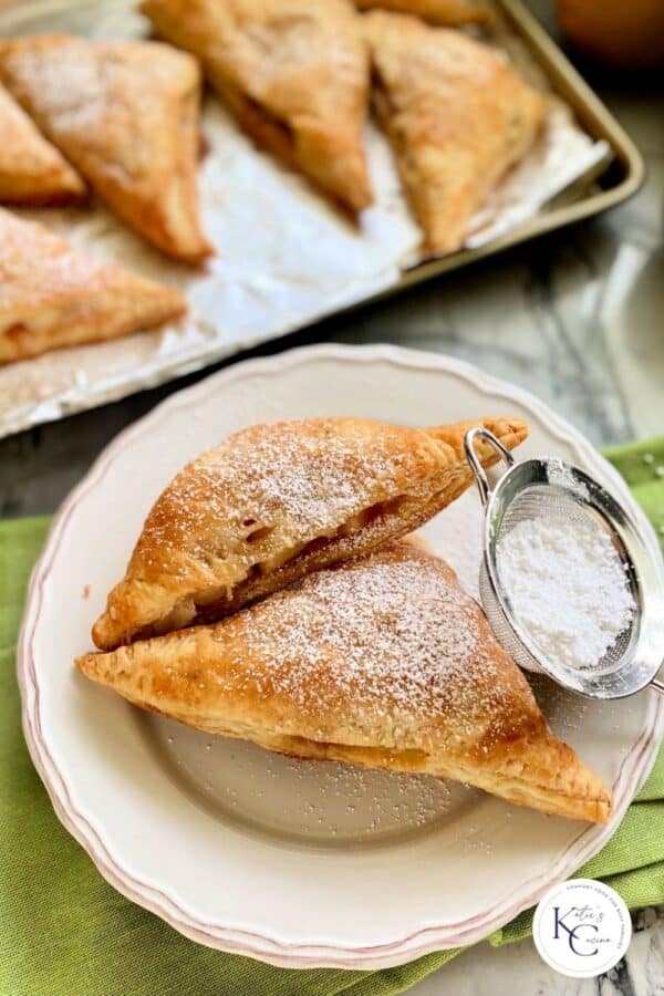 Two baked turnovers on a white plate with powder sugar sifter next to it with logo on bottom right corner.