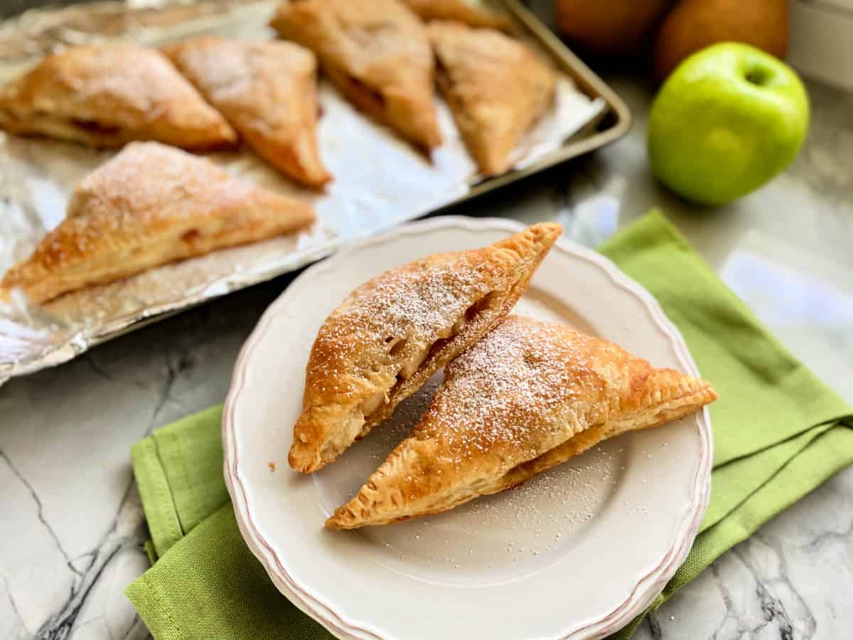 Two powder sugar dusted turnovers on a white plate with a tray of baked turnovers in the background. 