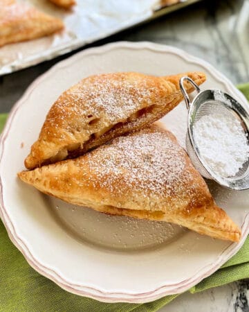 Two triangel turnovers on a white plate with a powder sugar sifter resting next to it.