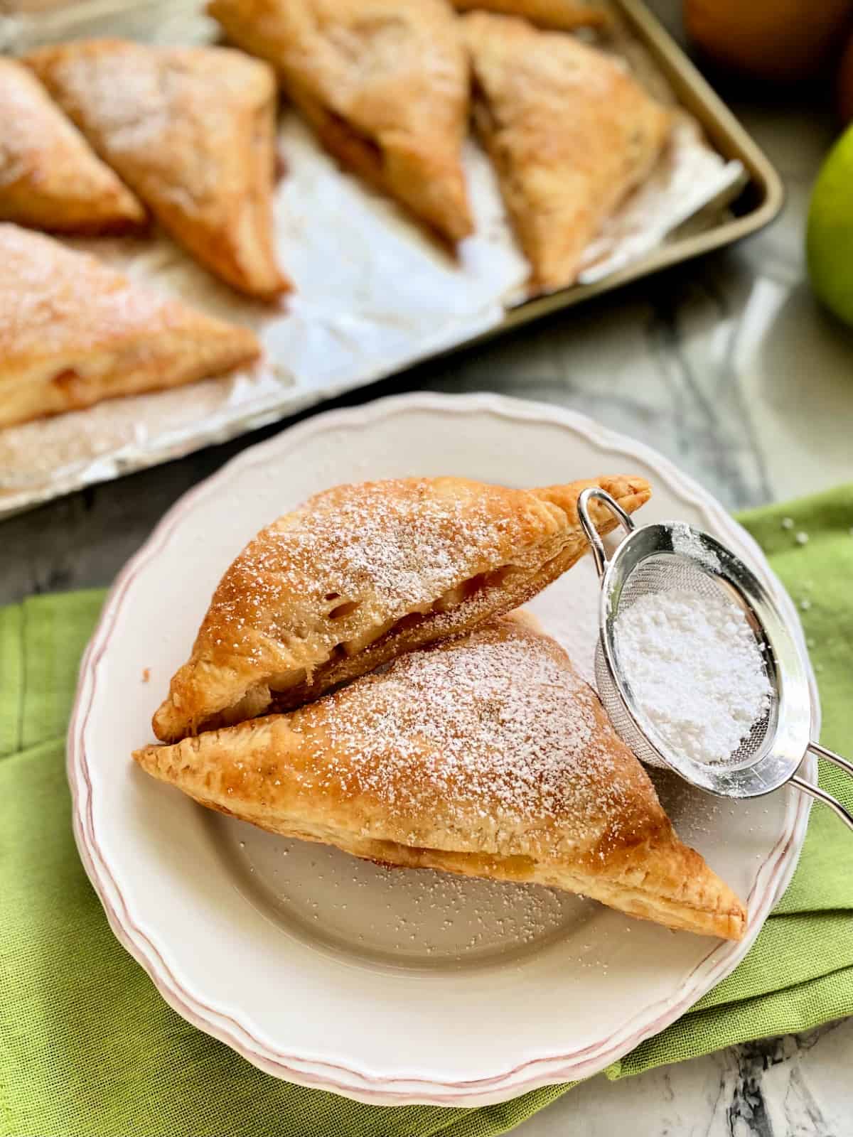 Top view of a white plate filled with apple turnovers and a mesh sifter filled with powder sugar.