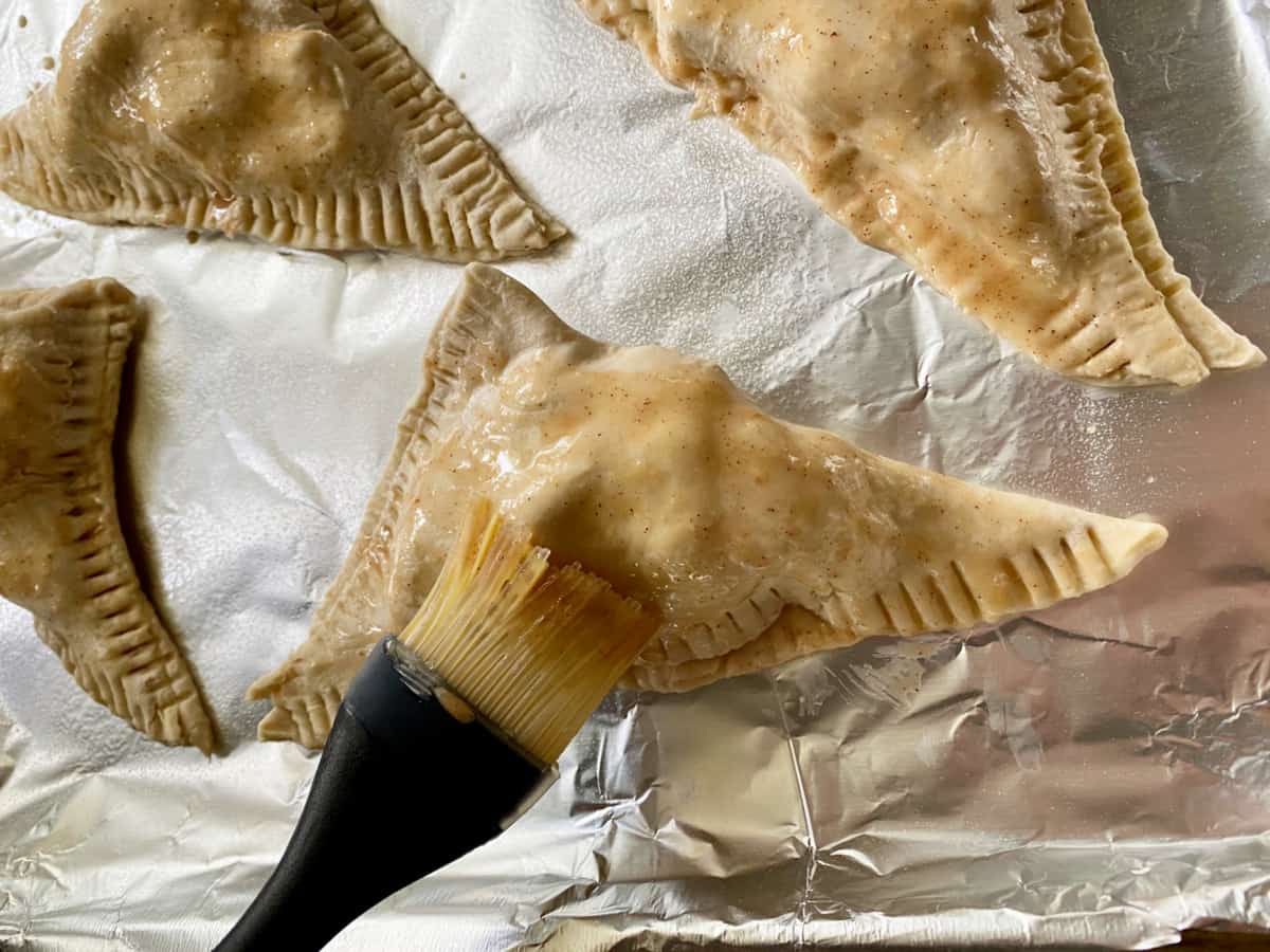 Silicone brush brushing cinnamon sugar mixture on top of raw puff pastry triangle. 