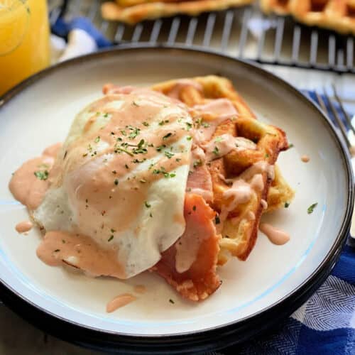 White plate filled with two waffles, ham, and an egg on top with cream sauce on top.