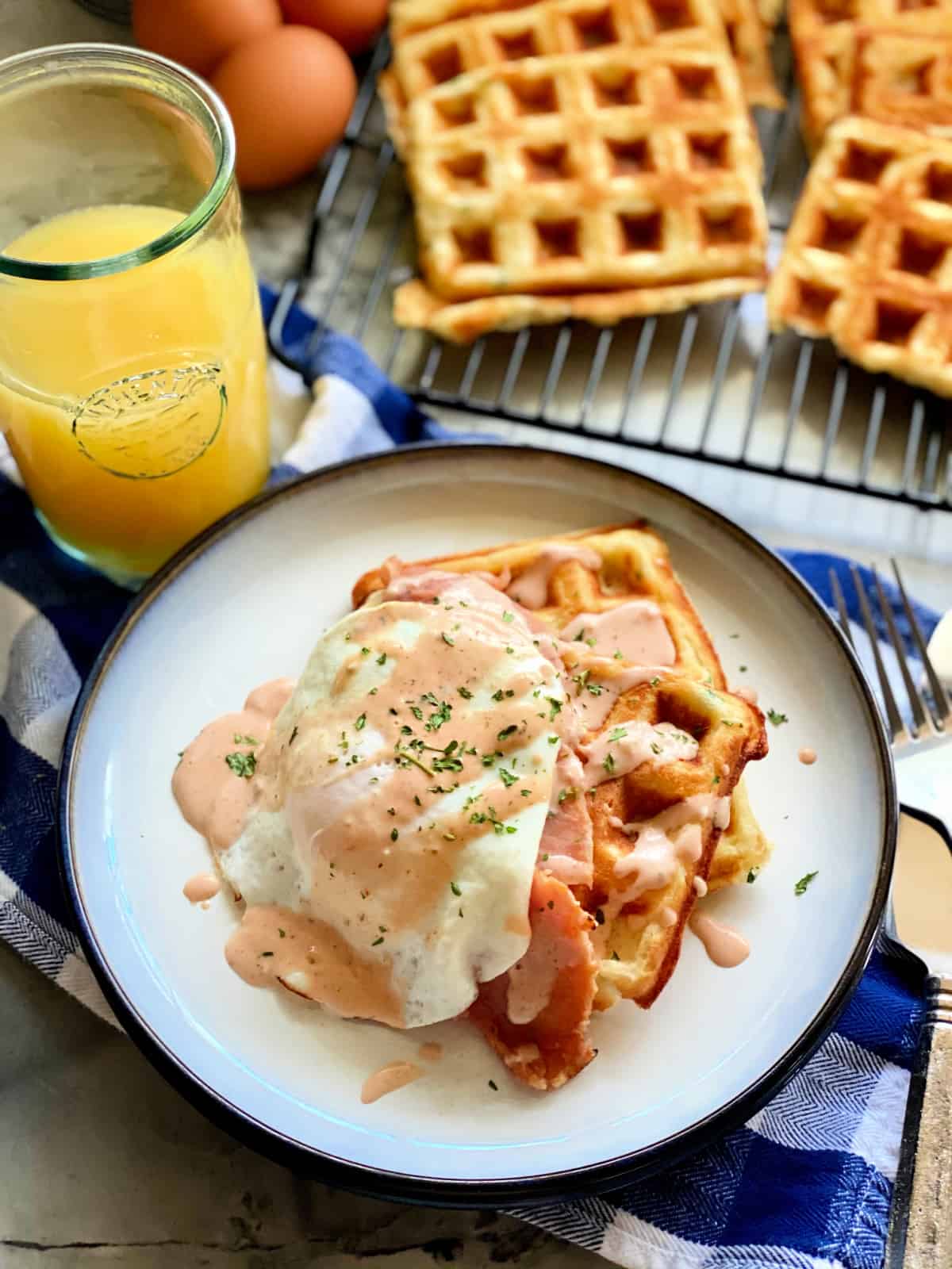 White plate with two waffles stacked with ham, egg, and sauce drizzled on top with logo on right bottom corner.