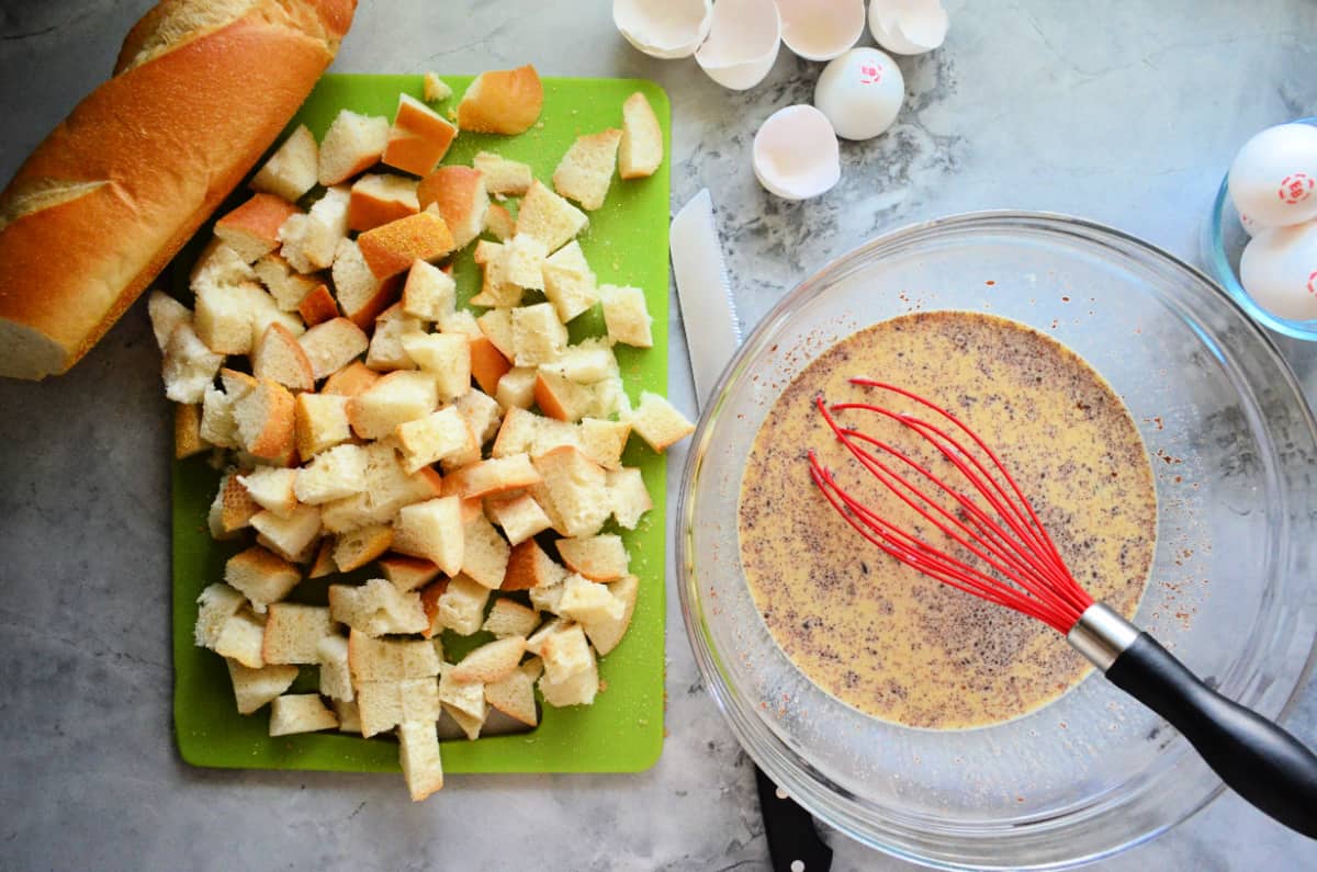 Top view of ingredients; left: baguette diced on cutting board, right: mixing bowl with egg mixture and whisk