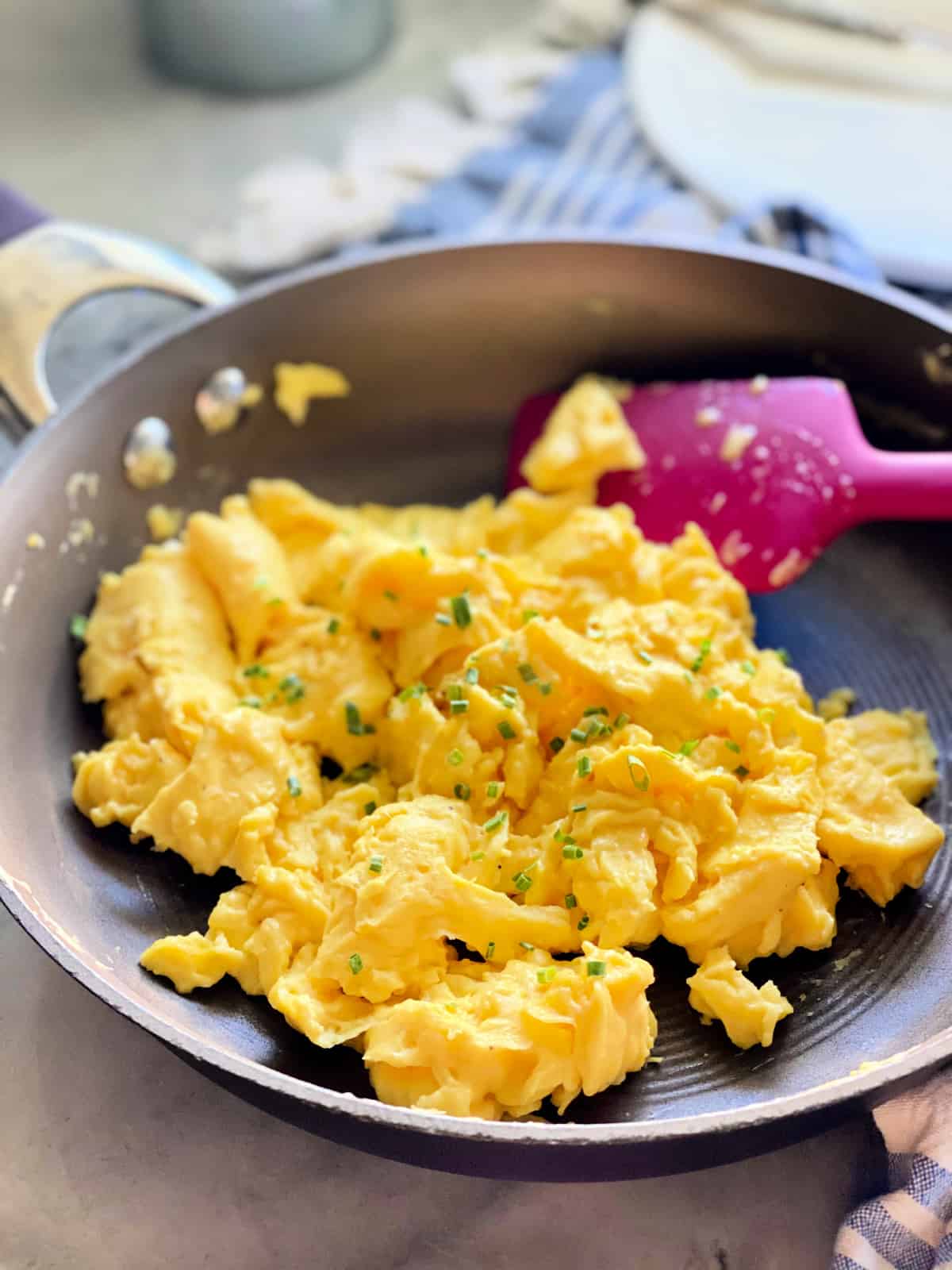 Brown frying pan with fluffy scrambled eggs topped with chives on a blue and white striped cloth.