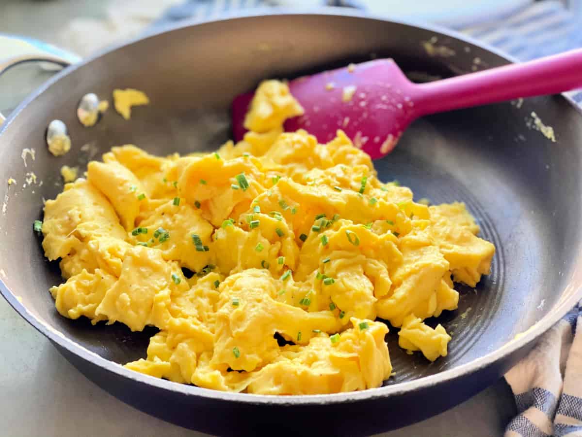 Brown frying pan with fluffy scrambled eggs topped with chives on a blue and white striped cloth.