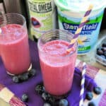 Two bright pink/purple smoothies on a purple cloth with blueberries around it and yogurt in the background.