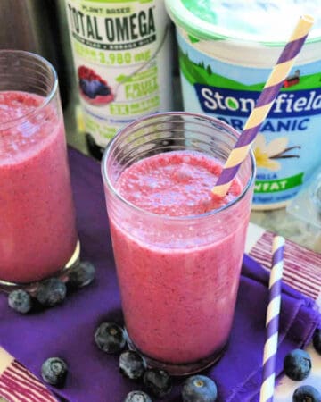 Two bright pink/purple smoothies on a purple cloth with blueberries around it and yogurt in the background.