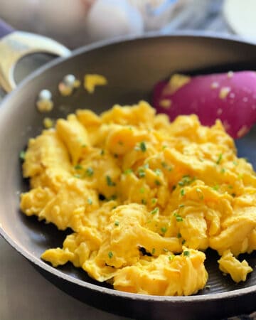 Brown skillet with scrambled eggs topped with chives and silicone spatula.