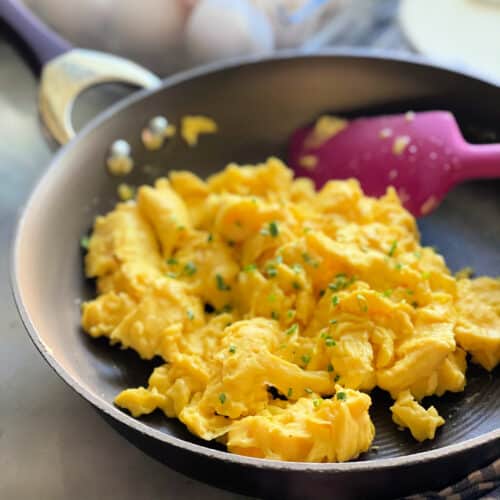 Brown skillet with scrambled eggs topped with chives and silicone spatula.