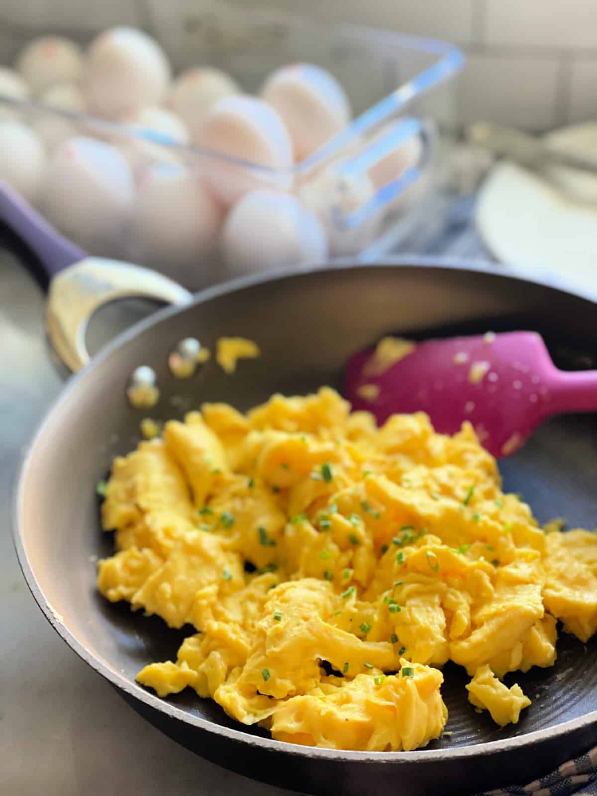 Brown frying pan with fluffy scrambled eggs topped with chives on a blue and white striped cloth