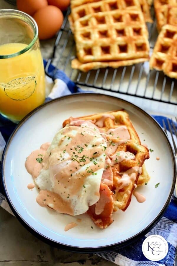 White plate with two waffles stacked with ham, egg, and sauce drizzled on top with logo on right bottom corner.