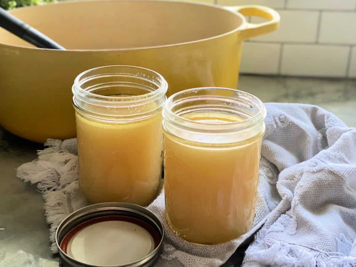 Two glass mason jars filled with chicken stock with a yellow pot in the background.