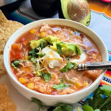 square photo of a white bowl filled with chicken soup, sour cream, cheese, and diced avocado with a spoon in the soup.