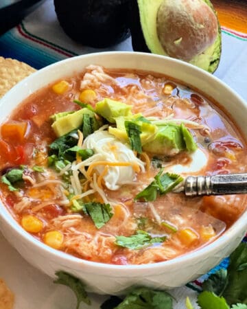 square photo of a white bowl filled with chicken soup, sour cream, cheese, and diced avocado with a spoon in the soup.