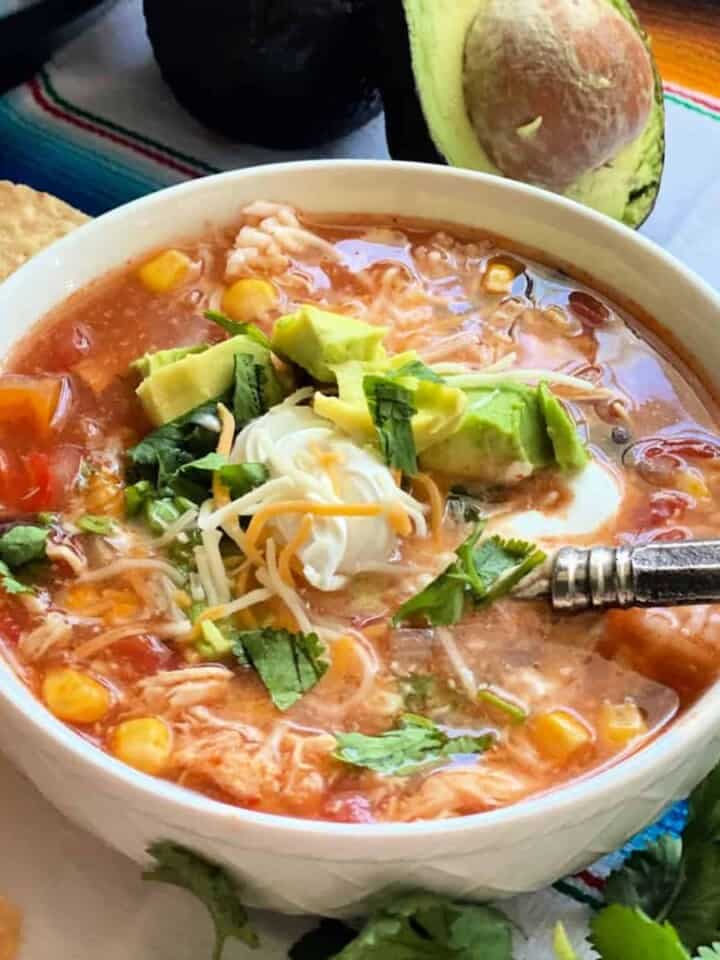 square photo of a white bowl filled with chicken soup, sour cream, cheese, and diced avocado with a spoon in the soup.