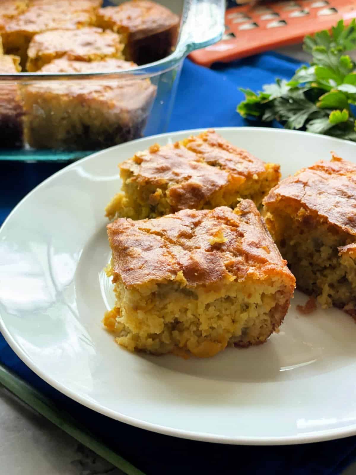 three cornbread slices on white plate with baking dish filled with more cornbread in the background