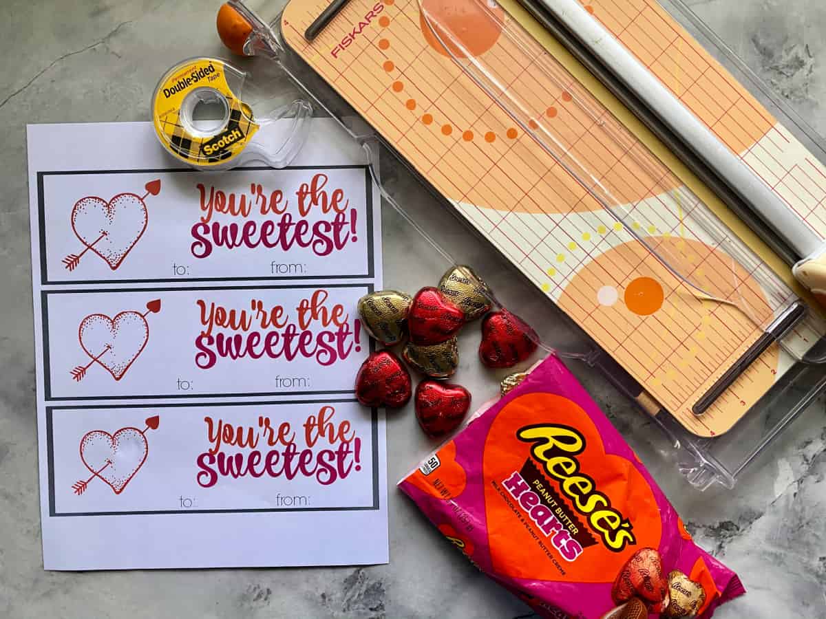 supplies on marble countertop: printable valentine, reese's chocolate hearts, double stick tape, and paper cutter.