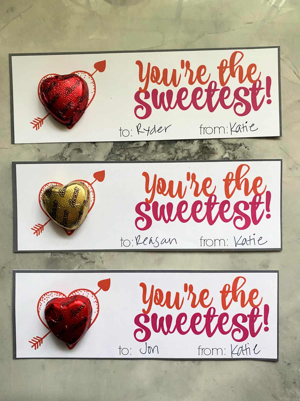 Three valentine's with heart chocolate candies resting on a marble countertop.