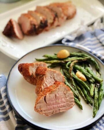 White plate with sliced pork tenderloins and green beans with pork tenderloin in the background.
