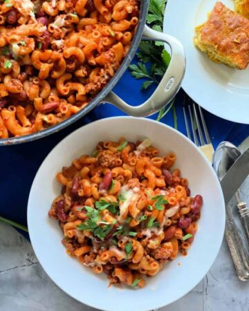 White bowl with pasta and ground beef with red sauce with green onions and parsley on top, pan of more pasta on the side.