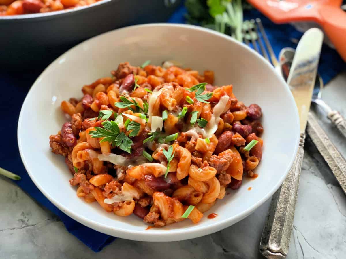 Chili with corkscrew pasta in white bowl with silverware next to it.