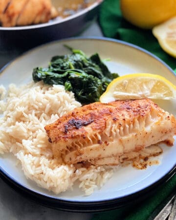 White plate with a piece of cod, white rice, spinach, and a lemon.