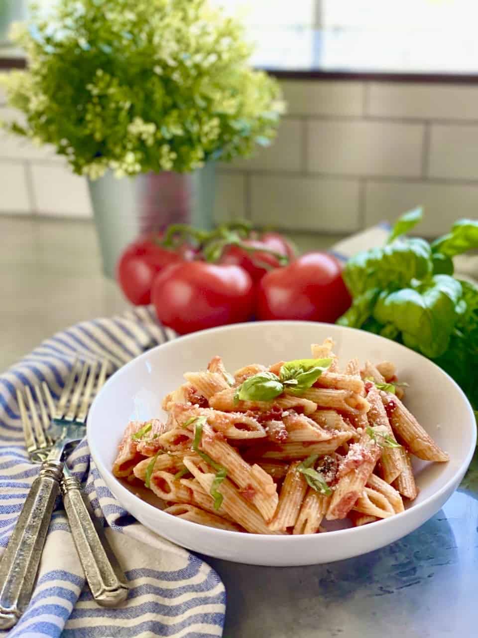 Penne Pomodoro in a white pasta plate with tomatoes and basil in the background