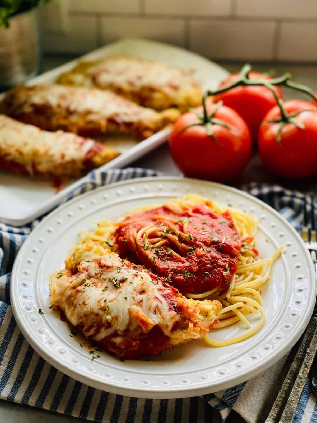 Chicken parm and spaghetti on a white plate with tomatoes on the vine and more chicken parm in the background