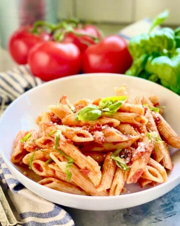 White bowl with penne pasta, tomato sauce, and basil with tomatoes in the background.