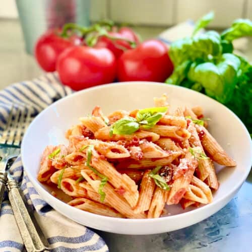 White bowl with penne pasta, tomato sauce, and basil with tomatoes in the background.