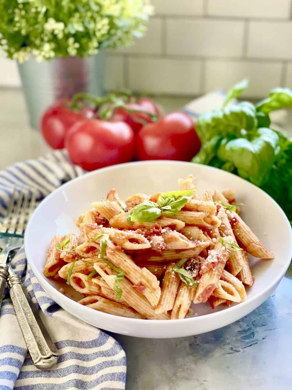 Penne Pomodoro in a white pasta plate with tomatoes and basil in the background