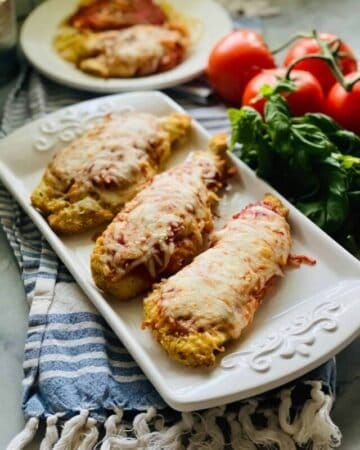white platter on a blue dish towel with three chicken parmesan breasts and a bowl of chicken parm and tomatoes in the background.