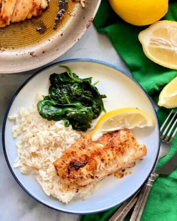 a white plate with a piece of blackened cod, rice, lemon slice, and spinach