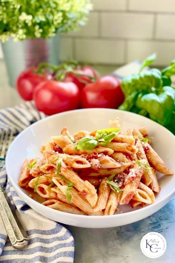 Penne Pomodoro in a white pasta plate with tomatoes and basil in the background