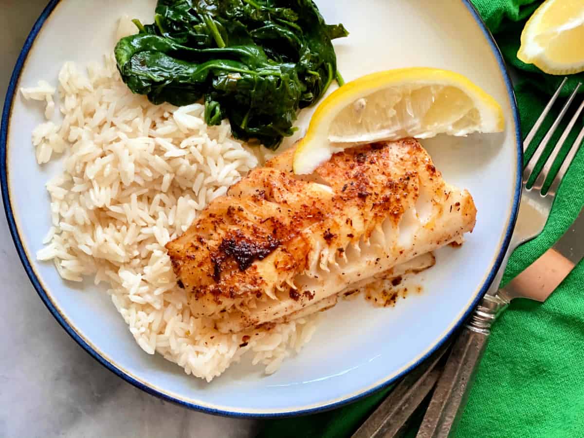 Top view shot of Blackened cod, white rice, spinach, and a lemon slice on a white plate