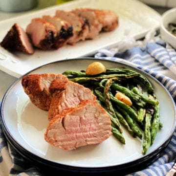 White plate with sliced pork tenderloins and green beans with pork tenderloin in the background.