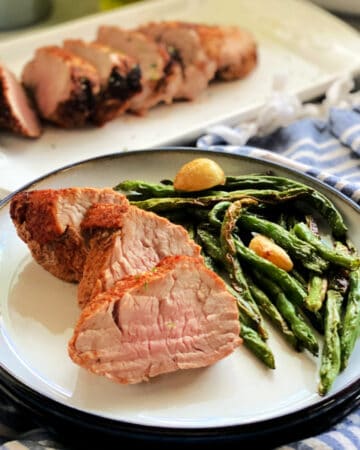 White plate with sliced pork tenderloins and green beans with pork tenderloin in the background.