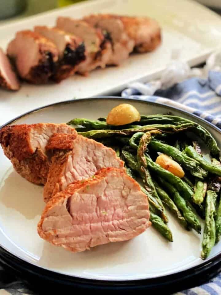 White plate with sliced pork tenderloins and green beans with pork tenderloin in the background.