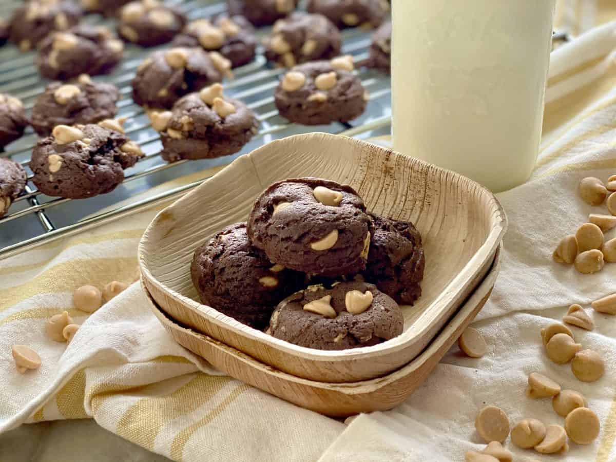 Four chocolate peanut butter chip cookies in a wooden bowl with more cookies on a wire rack in the background