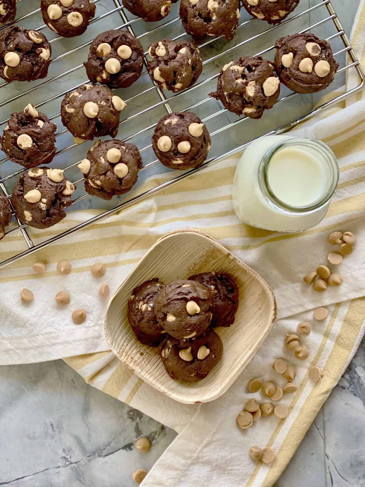 Top view shot of Four chocolate peanut butter chip cookies in a wooden bowl with more cookies on a wire rack and glass of milk in the background