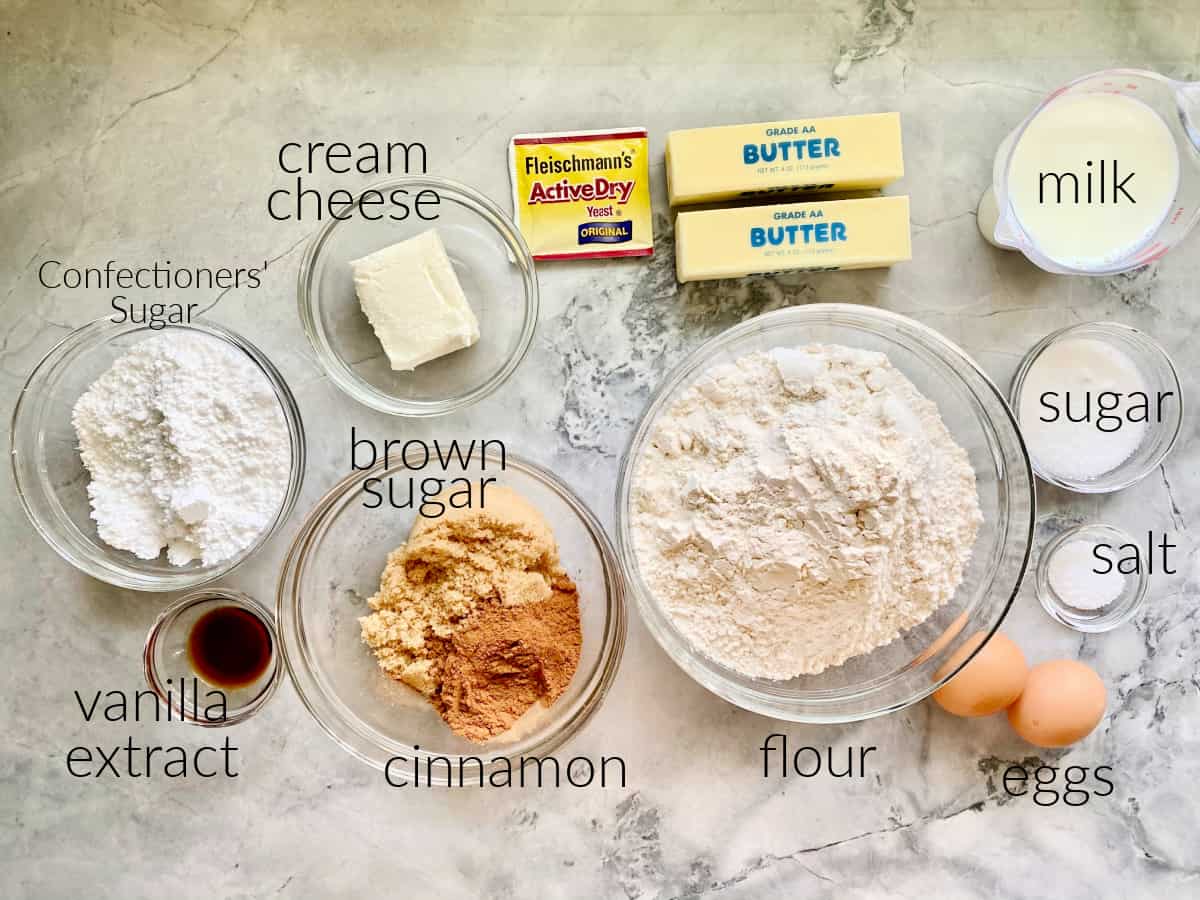 Ingredients on marble countertop: sugar, vanilla, cream cheese, egg, cinnamon, yesat, butter, milk, flour, and salt.