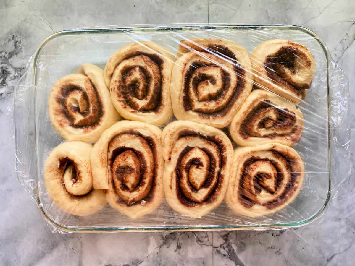 Glass baking dish with cinnamon rolls in it covered with plastic wrap.