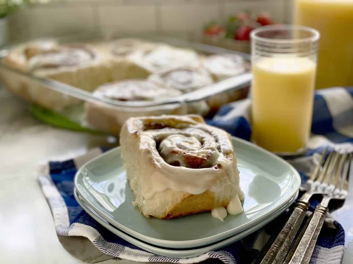 Cinnamon roll on blue square plates with tray of cinnamon rolls in the background.