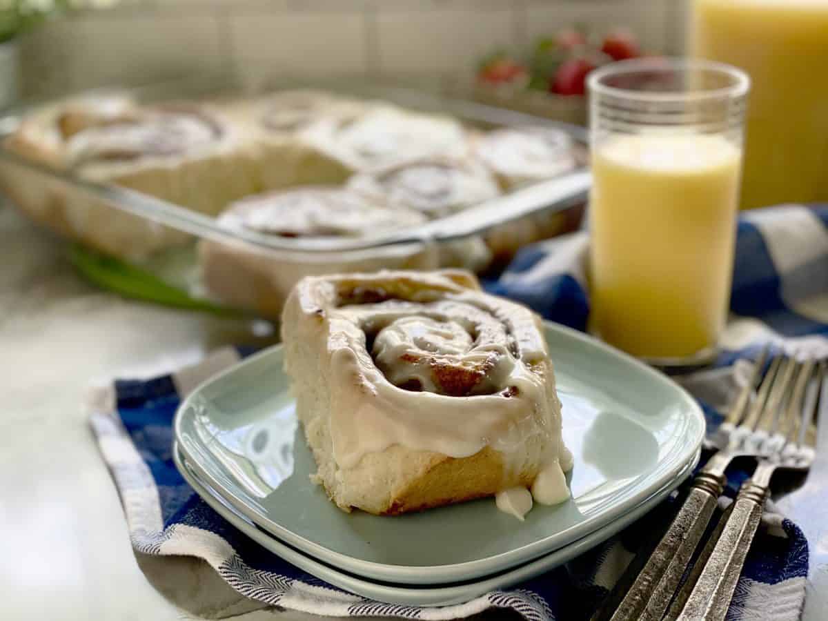 Homemade Cinna-Buns from katiescucina.com Cinnamon roll on blue square plates with tray of cinnamon rolls in the background.