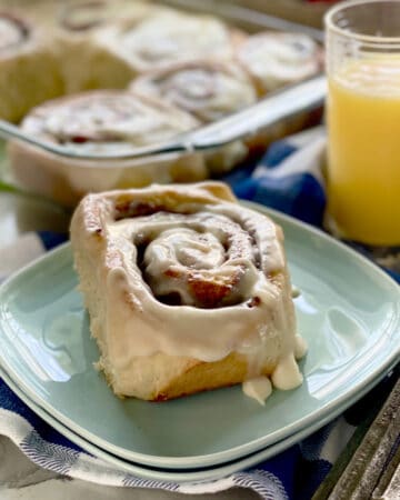 Two blue plates stacked with a thick cinnamon roll on top with orange juice in background.