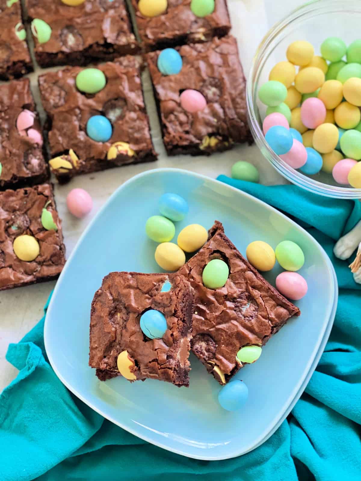 Top view of two blue plates with two brownies and brownies cut and a bowl of candy eggs on the side.