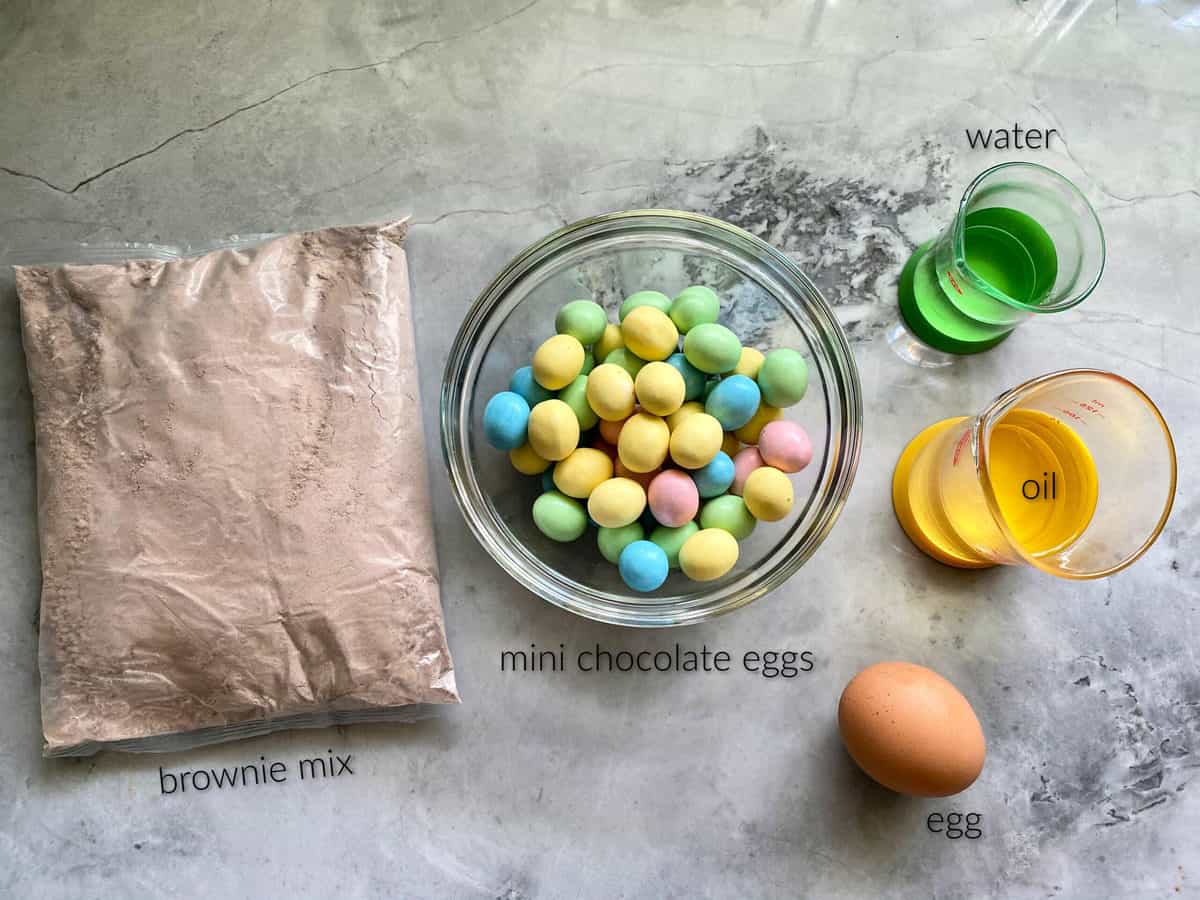Ingredients on marble counter; brownie mix, mini chocolate eggs, water, oil, and egg.